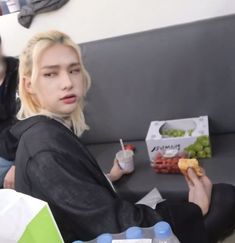 a person sitting on a couch holding a drink and eating food with a box of fruit in front of them