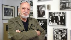 an older man with his arms crossed standing in front of a wall full of black and white photos