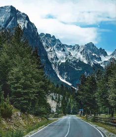 an empty road in the mountains with snow on the top and green trees around it