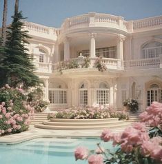 a large white house with pink flowers in the foreground and a pool surrounded by trees