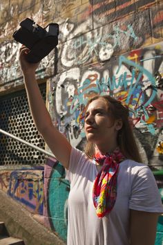 a woman holding up a cell phone in front of a graffiti covered wall with stairs