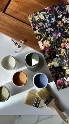 paint and brushes on a table with floral wallpaper