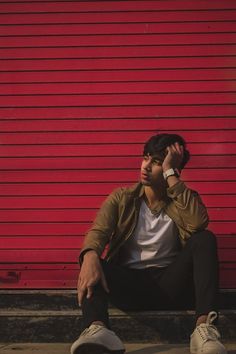 a man sitting against a red wall talking on a cell phone while holding his hand up to his ear