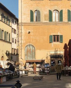 people are walking around an outdoor market in front of a building with green shutters