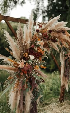 an arrangement of dried flowers and feathers is displayed in front of a wooden structure on the grass