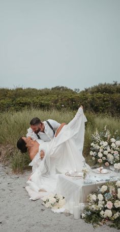 a bride and groom kissing in front of their wedding cake on the beach with flowers