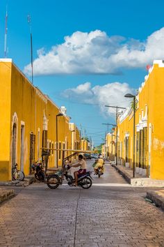 a man riding a motorcycle down a street next to tall yellow buildings on either side