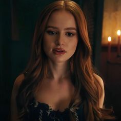 a woman with long red hair standing in front of a candle lit room and looking at the camera