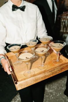 a man in a tuxedo holding a tray with four martini glasses on it