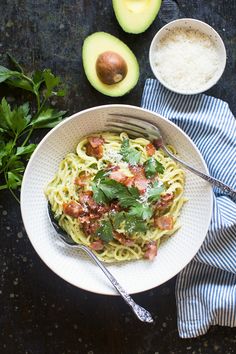 a bowl of pasta with bacon, parsley and avocado on the side