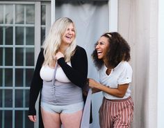 two women standing next to each other in short shorts and high waisted panties, laughing