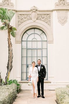 a man and woman standing in front of a building with an arched door, dressed in formal attire