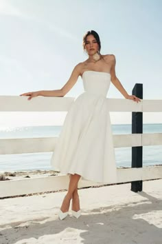 a woman in a white dress leaning on a fence by the beach with her arms outstretched