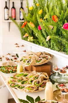 an assortment of food is displayed on a buffet table in front of plants and wine bottles