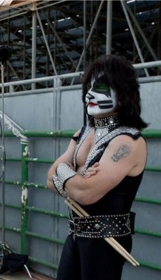 a man in black and white makeup is standing with his arms crossed while holding two drums