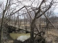 an old bridge over a small stream in the middle of trees and grass with no leaves on it