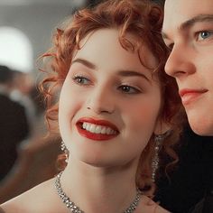 a woman with red hair and a man in black are looking into each other's eyes