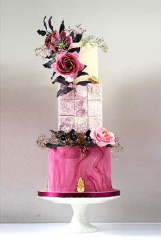 a three tiered cake decorated with pink and purple flowers on a white pedestal in front of a gray wall