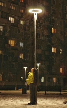 a woman standing next to a street light in the snow at night with buildings behind her