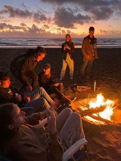 group of people sitting around a campfire on the beach at sunset with one person standing