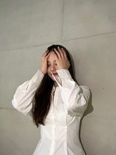 a woman is covering her face with her hands while standing against a wall and looking at the camera