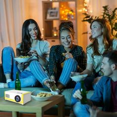 three women and two men are sitting on the couch eating food, drinking beer and watching tv