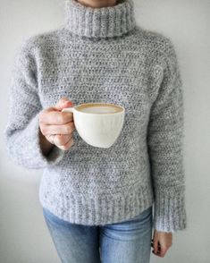 a woman holding a coffee cup in her right hand and wearing a gray knitted sweater