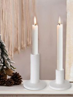 two white candles sitting on top of a table next to a pine cone and christmas tree