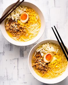 two bowls filled with noodles, meat and eggs on top of a white wooden table