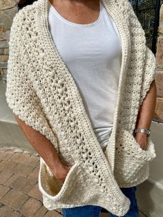 a woman wearing a white crocheted cardigan and jeans standing in front of a brick wall