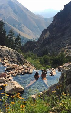 two people in a hot tub surrounded by rocks and flowers with mountains in the background