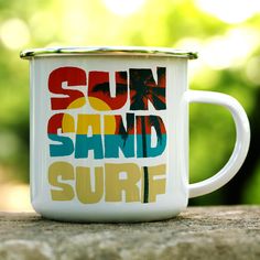 a white coffee mug with the words sun sand surf on it sitting on a rock