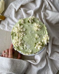 a person holding a green cake with white flowers on it and napkins next to it