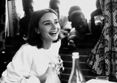 a black and white photo of a woman laughing while sitting at a table with other people in the background