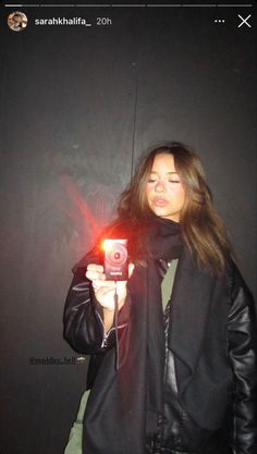 a woman taking a selfie with her camera in front of a black wall and wearing a leather jacket