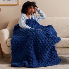 a woman is sitting on a couch wrapped in a blue knitted blanket and looking at the camera