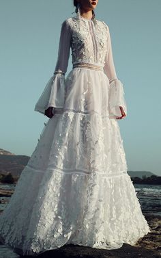 a woman is standing on the beach wearing a white dress with ruffles and bell sleeves