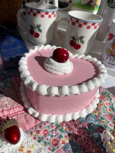 there is a pink cake with white frosting and a cherry on top, next to two coffee mugs