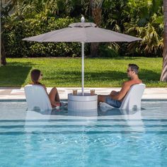 two people sitting in chairs under an umbrella next to a swimming pool with grass and palm trees