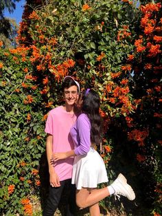 two young people standing next to each other in front of orange and red flowers on a bush