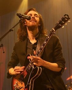 a man with long hair holding a guitar and singing into a microphone while standing in front of two microphones