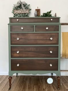 an old dresser painted green and brown with white knobs on the top, sitting in front of a wall