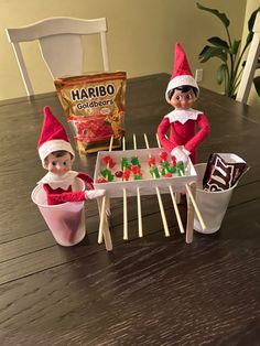 two elfs sitting at a table with candy and marshmallows on sticks