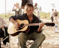 a man sitting in a chair with a guitar and people around him playing music on the beach