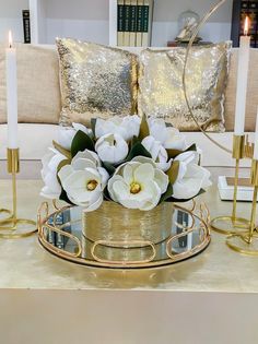 white flowers in a gold vase on a glass tray with candles and pillows behind it