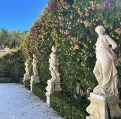 there are many statues on the side of this wall that is lined with bushes and flowers