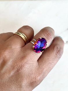 This stunning crystal cocktail ring is sure to turn heads! A gorgeous 18mm oval crystal is set in a luxurious, adjustable gold plated ring band that can fit sizes 5 and up. This 12-carat gem is perfect for any special occasion or for adding an eye-catching sparkle to your everyday look. As the light catches it, the crystal gleams in a shades of blue, purple, and pink. The ring base is nickel free! Ships Free • Handmade in the USA Purple Oval Amethyst Crystal Ring, Unique Adjustable Purple Crystal Ring, Purple Round Crystal Spiritual Ring, Purple Spiritual Crystal Ring, Oval, Elegant Purple Multi-stone Crystal Ring, Ring Sale, Oval Rings, Gold Plated Rings, Pink Crystal