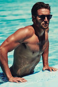 a shirtless man in the water on a surfboard wearing sunglasses and swimming trunks