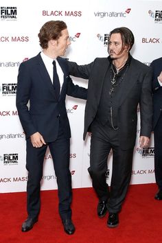 two men in suits standing on a red carpet with one pointing at the other man