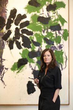 a woman standing in front of a painting holding a paintbrush and looking at the camera